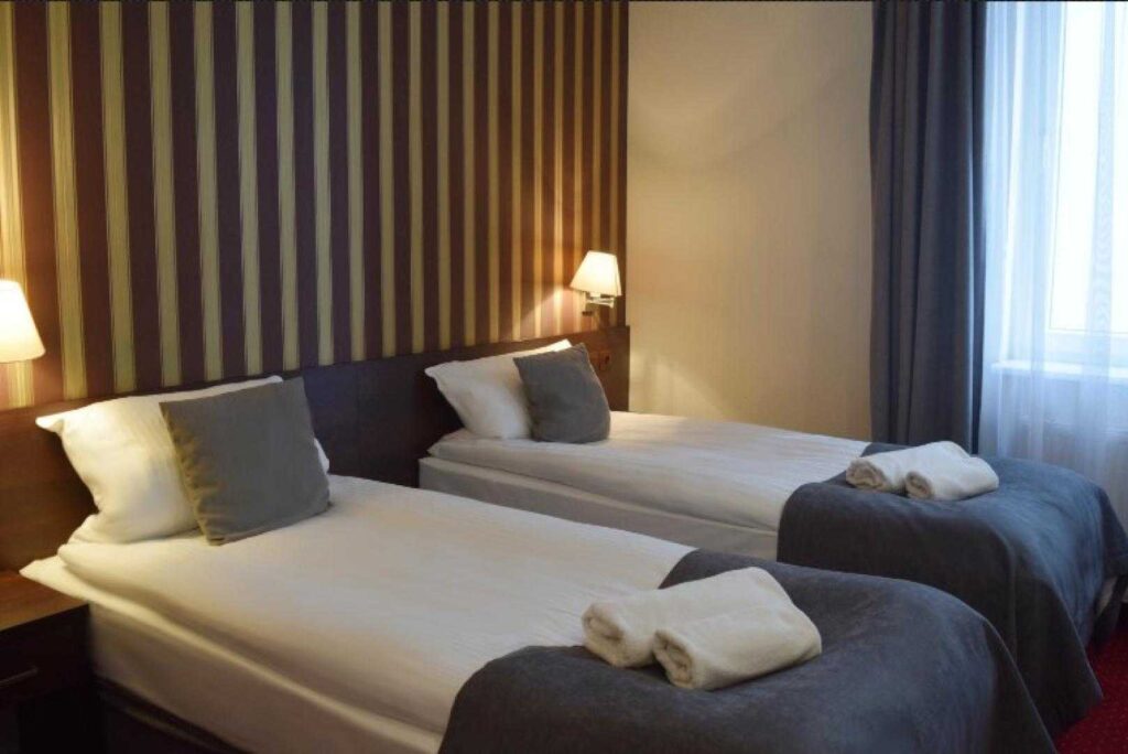 Dwuosobowy pokój w hotelu Maraton z dwoma oddzielnymi łóżkami, szarymi narzutami i pasiastą tapetą.